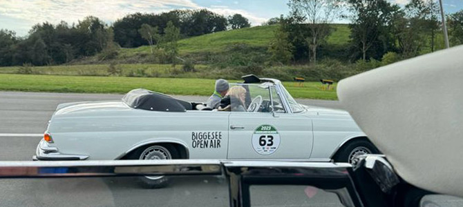 Ein weißes Oldtimer-Cabrio mit einem Biggesee Open Air-Schild und einem Aufkleber mit der Nummer 63, das von zwei Personen gefahren wird, befindet sich auf einer Straße mit grasbewachsenen Hügeln im Hintergrund.
