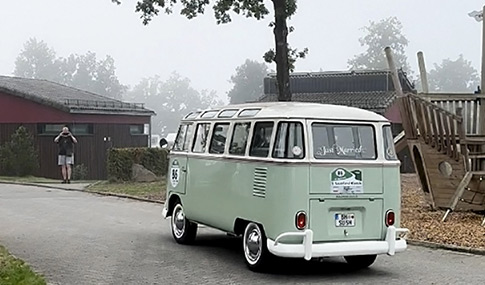 Ein alter hellgrüner Volkswagen Naturwagen fährt auf einer nebligen Straße in der Nähe eines Spielplatzes, während im Hintergrund eine Person ein Foto macht.
