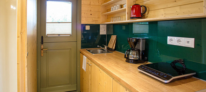 Compacte Naturwagen keuken met houten kastjes, een groene deur, gootsteen, koffiezetapparaat, waterkoker, inductiekookplaat en planken met borden en kopjes.