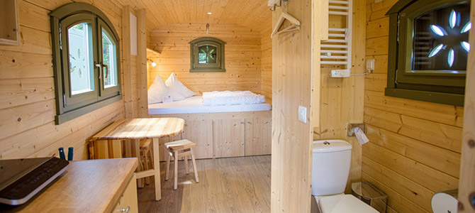 Interieur van een kleine Naturwagenhut met een bed, tafel met krukje, toilet en ramen met groene rand; alle meubels en muren zijn gemaakt van licht hout.