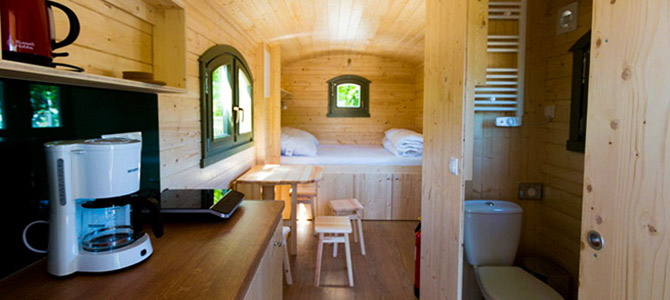 Interieur van een gezellige Naturwagen met een kleine houten cabine met een keukentje, koffiezetapparaat, eettafel, netjes opgevouwen beddengoed en een compacte badkamer met toilet zichtbaar.