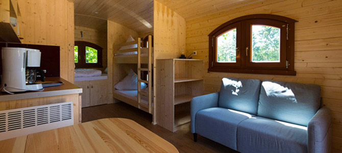 Interieur van een kleine houten Naturwagen met een blauwe bank, stapelbed, tafel, raam en een keukentje met koffiezetapparaat.