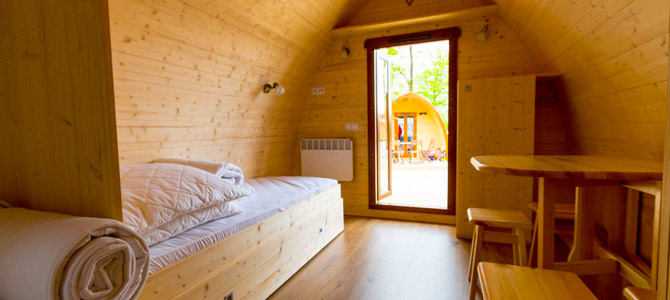 Interieur van een kleine houten hut met een eenpersoonsbed, opgevouwen beddengoed, een tafel met krukjes en een open deur die een andere soortgelijke hut buiten laat zien.