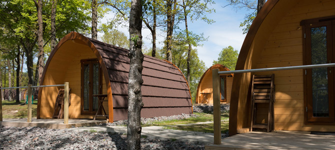 De houten Naturlodge-campings met gewelfde daken liggen verscholen tussen de bomen, elk met een kleine veranda en grindpaden, allemaal badend in de zon.