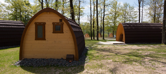 Verschillende kleine, houten hutten in de vorm van een Naturlodge pod staan tussen de bomen op een grasveld onder een heldere hemel.