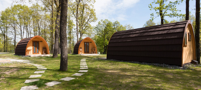 Drie Naturlodge houten hut pods met gebogen daken rusten op een grasveld met stapstenen paden, omgeven door bomen onder een gedeeltelijk bewolkte hemel.