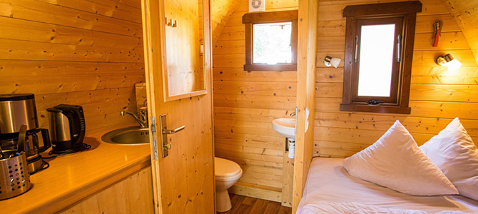 Kleine houten hut met een kitchenette, gootsteen, waterkoker en koffiezetapparaat aan de linkerkant, een bed met kussens aan de rechterkant en een compacte badkamer zichtbaar door een open deur.