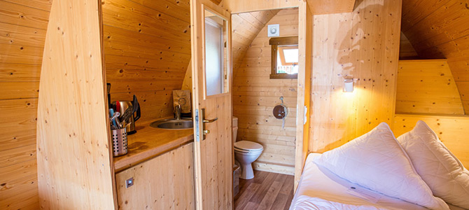 Interieur van een kleine houten hut met een keukentje, gootsteen, keukengerei en een aparte ruimte met een toilet. Een bed met witte kussens is zichtbaar op de voorgrond.