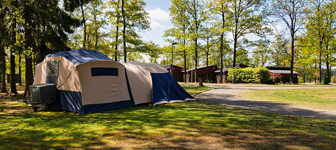 Een grote tent staat op gras op een beboste camping, met een verhard pad en rode gebouwen zichtbaar op de achtergrond op een zonnige dag.