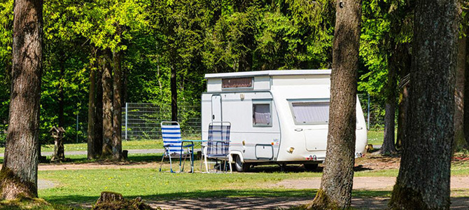 Een witte camper staat geparkeerd op gras naast bomen, met twee gestreepte tuinstoelen en een tafel buiten. Op de achtergrond zijn bos en een draadomheining zichtbaar.