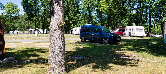 Een blauw busje staat geparkeerd op gras bij een boom aan de Rondell, met verschillende witte caravans en aanhangers op de achtergrond tussen hoge groene bomen.