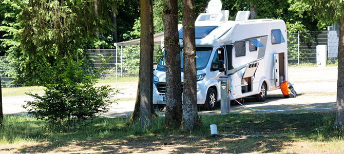 Een witte camper staat geparkeerd op een camping tussen de bomen, verbonden met een Komfortplatz Säule met kabels en slangen op een zonnige dag.