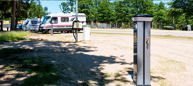 Campers geparkeerd op een terrein met Komfortplatz Säule elektrische aansluitpunten en bomen op de achtergrond onder een heldere hemel.