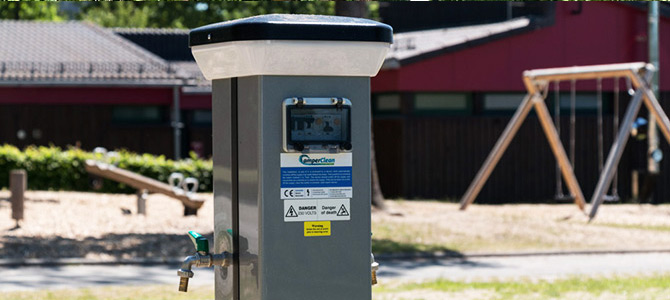 Een Komfortplatz Säule waterdistributiestation met kraantjes wordt geïnstalleerd in een buitenruimte in de buurt van een speeltuin met schommels en andere speelstructuren.