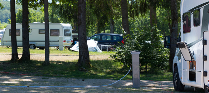 Camping Komfortplatz met meerdere geparkeerde caravans, een auto en een tent tussen de bomen op een zonnige dag; stroomaansluiting zichtbaar op de voorgrond.